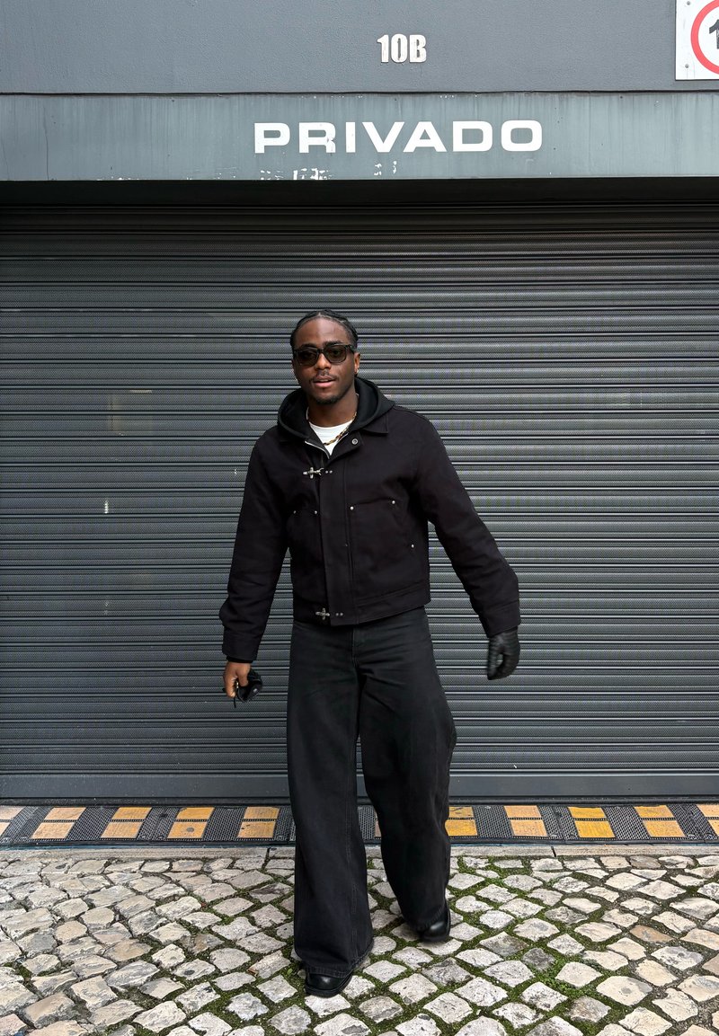 Mees mustade päikeseprillide, jaku, kinnaste ja laiate pükstega kõnnib munakiviteel halli rullukse ees, mille kohal on tekst "PRIVADO".