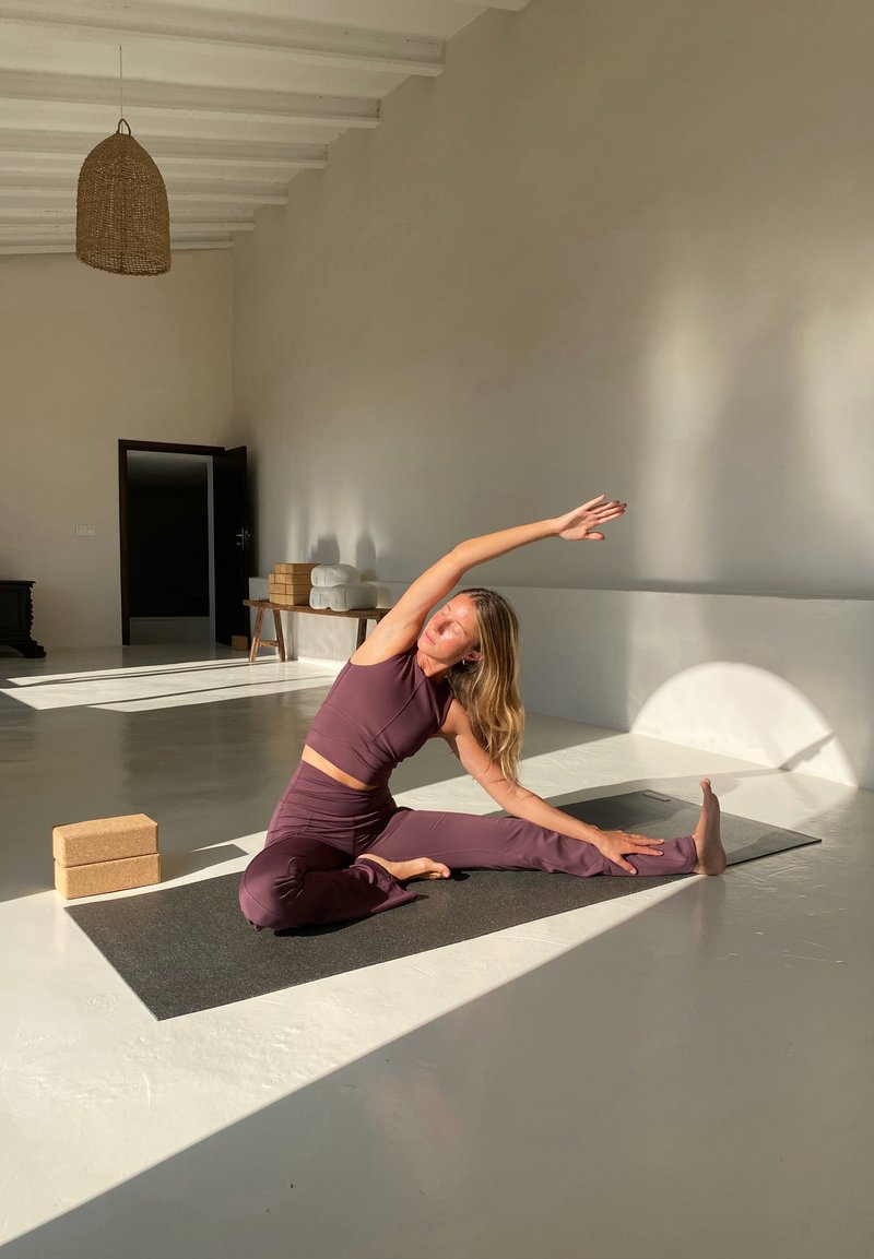 Femme en tenue de sport violette s'étirant sur le côté sur un tapis de yoga dans une pièce lumineuse et spacieuse avec des blocs de yoga à proximité.