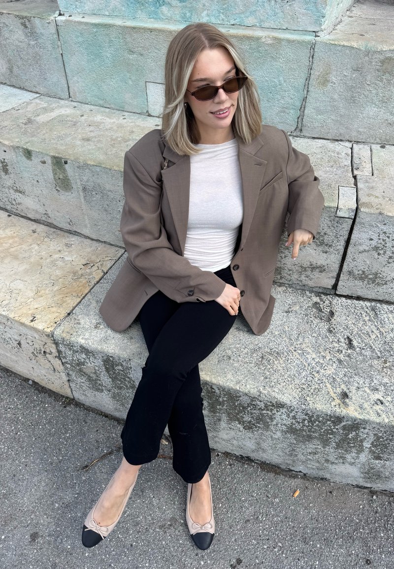 Jeune femme avec des lunettes de soleil, un blazer beige, un haut blanc et un pantalon noir assise sur des marches en pierre à l'extérieur.