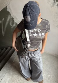 Chemise à manches courtes camouflage grise avec texte blanc "NYC 90", associée à un pantalon gris ample. Casquette noire avec logo blanc et sac à main noir.