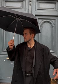 Svart paraply hålls av en man klädd i svart frack och skjorta, stående mot en strukturerad ljusblå dörr med dekorativa paneler.