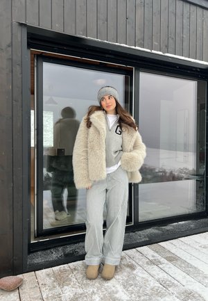 Abrigo de piel sintética color crema gruesa, suéter de punto gris con una 'G', camisa blanca, pantalones a rayas gris y blanca, y botas de tobillo beige. Fondo nevado.