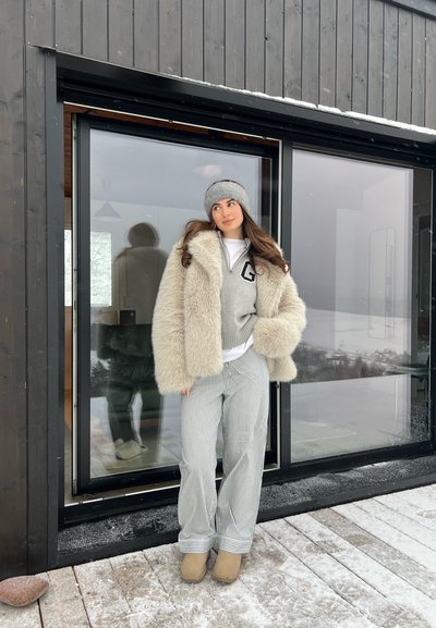 Abrigo de piel sintética color crema gruesa, suéter de punto gris con una 'G', camisa blanca, pantalones a rayas gris y blanca, y botas de tobillo beige. Fondo nevado.