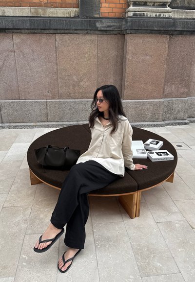 Banco de madera curvado con tapicería de color marrón oscuro, que presenta una superficie lisa. A su lado hay un bolso negro y libros apilados.