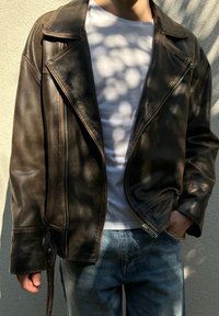 Brązowa skórzana kurtka motocyklowa z wyciętym kołnierzem, asymetrycznym zamkiem błyskawicznym i bocznymi kieszeniami, noszona na białej koszulce i niebieskich dżinsach.