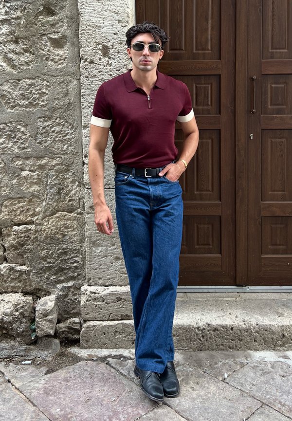Hombre con gafas de sol, una camiseta tipo polo burdeos, vaqueros azules y zapatos negros, de pie sobre un pavimento de piedra junto a una puerta de madera y una pared de piedra.