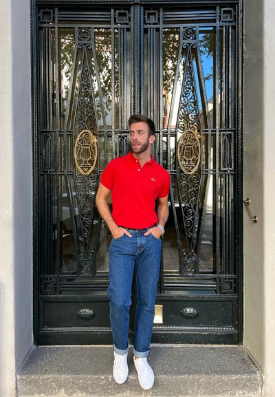 Camisa polo roja, jeans azules y zapatillas blancas contra puertas de metal negro ornamentadas con acentos dorados y detalles decorativos.