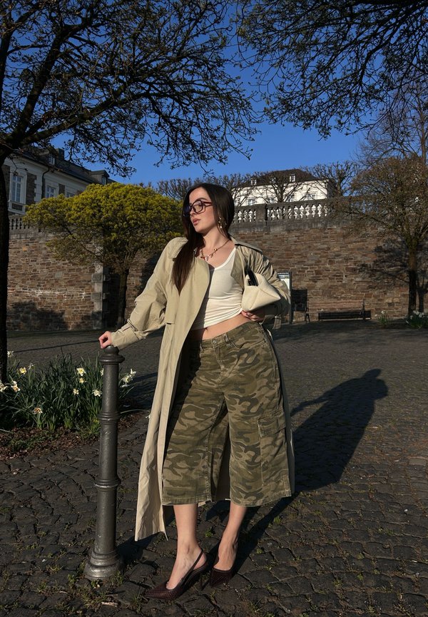 Cappotto trench beige, top bianco attillato, pantaloni a gamba larga con motivo camouflage, scarpe col tacco bordeaux, in piedi accanto a un palo di metallo.