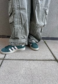 Green suede sneakers with white stripes and textured rubber soles, paired with loose, textured gray cargo pants, resting on a paved surface.