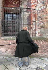 Un homme portant un long manteau sombre et des lunettes de soleil se tient face à une fenêtre grillagée installée dans un mur de briques et de pierres usées par le temps.