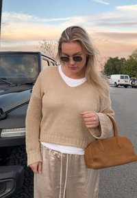 Femme portant un pull en tricot beige et des lunettes de soleil, debout à côt é d'un SUV noir, tenant un sac à main marron dans la rue au coucher du soleil.