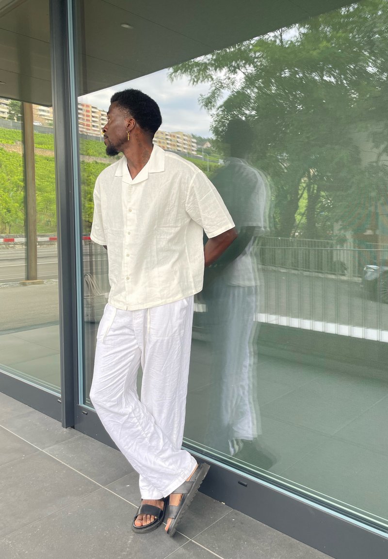 Chemise blanche à manches courtes avec boutons et pantalon blanc léger, assortis à des sandales noires, se tenant devant un mur de verre reflétant de la verdure.