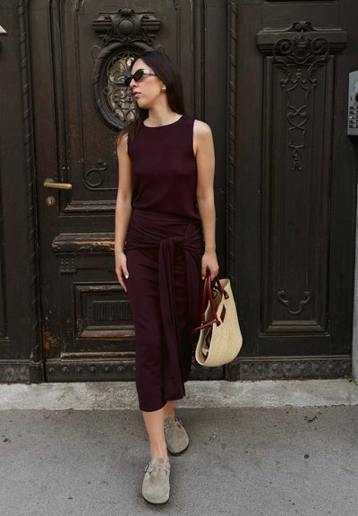 Top sin mangas burdeos con un detalle anudado, falda midi a juego, zapatos grises y un bolso de paja con asas rojas, todo dispuesto frente a una puerta oscura.