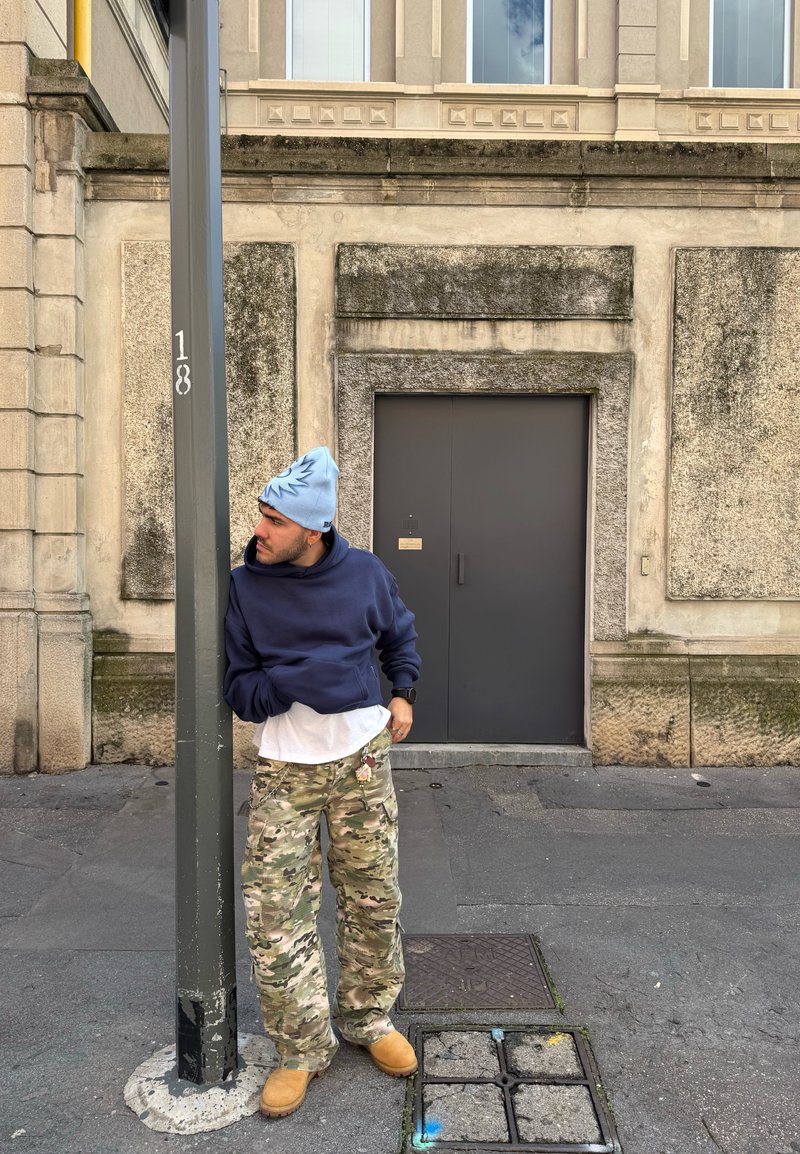 Αρκετά casual ντύσιμο με ένα μπλε hoodie, λευκό πουκάμισο, καμουφλάζ cargo παντελόνια και μπεζ μπότες. Μπλε σκουφί με σχέδιο ήλιου. Στηρίζεται σε έναν στύλο.