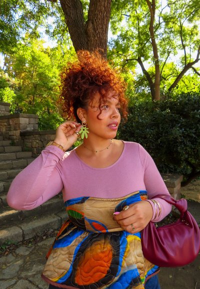 Mujer con pelo rizado rojo ajustando un pendiente verde en forma de flor, vistiendo una blusa rosa y un chaleco acolchado colorido, sosteniendo un bolso burdeos al aire libre.