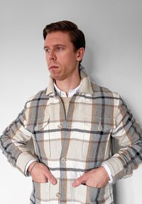 Homme aux cheveux bruns courts portant une veste  à carreaux beige, blanche et noire, mains dans les poches, regardant pensivement vers la gauche sur un fond gris uni.