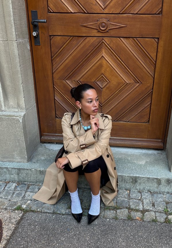 Femme en trench beige, leggings noirs, chaussettes blanches et chaussures noires assise sur des marches en pierre devant une porte en bois sculpté, regardant vers la droite.