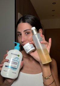 Drie huidverzorgingsproducten vastgehouden: een witte cleanser met pompdispenser, gelabeld "Hyaluronic Barrier Pro," een witte crème pot, en een heldere, frosted vloeistoffles met een gouden dop.