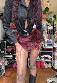 Mini saia de pelúcia cor de vinho com uma textura irregular e fofa. Usada com uma camisa preta com botões, abotoada abaixo da cintura. Botas pretas até o joelho.