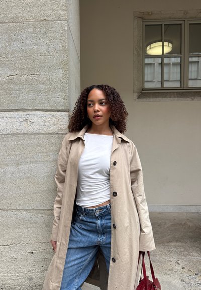 Mujer con cabello rizado que lleva un abrigo beige, camisa blanca y vaqueros azules, de pie junto a una pared de piedra cerca de una ventana.