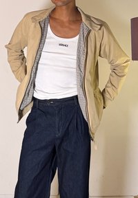 Άνδρας που φοράει μπεζ μπουφάν πάνω από λευκό τοπ Versace χωρίς μανίκια και σκούρο μπλε ψηλόμεσο παντελόνι, στέκεται με τα χέρια πίσω από την πλάτη.