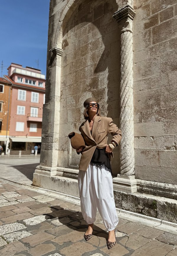Woman wearing brown blazer, beret, sunglasses, white loose trousers and leopard print shoes stands by ornate stone arch in sunny urban square.