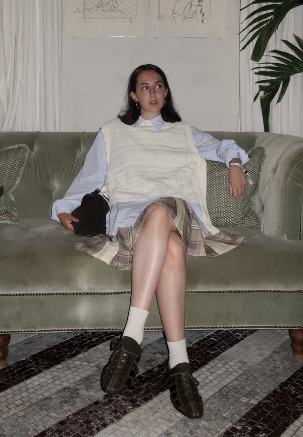 Femme aux cheveux foncés assise sur un canapé vert portant un gilet en laine blanc, une jupe à carreaux, une chemise blanche, des chaussettes et des chaussures marron, tenant un sac noir.