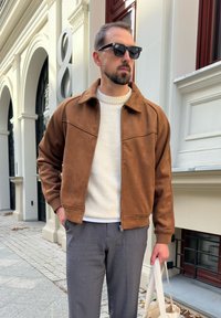 Chaqueta de ante marrón con cuello, bolsillos frontales y cierre de cremallera, llevada sobre un suéter blanco y pantalones grises. Gafas de sol negras.