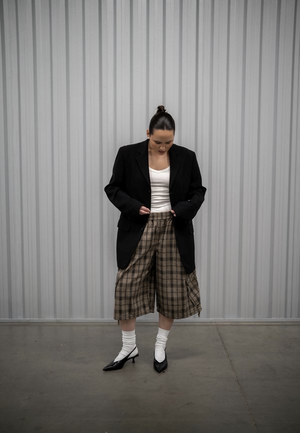 Femme en blazer noir, haut blanc, pantalon large à carreaux, chaussettes blanches et talons noirs, debout contre un mur métallique gris ondulé, regardant vers le bas.