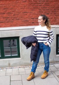 Top de rayas blancas y azul marino de manga larga, jeans de tiro alto azules, chaqueta acolchada negra y botas de cordones color marrón claro sobre una superficie pavimentada.