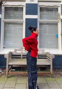 Maglione a collo alto a coste rosse, collo alto, vestibilità ampia, abbinato a jeans in denim scuro con cuciture in vista e tasche, posizionato davanti a un muro blu.