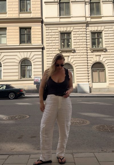 Top de tirantes negro con volantes, pantalones cargo de lino blancos, chanclas negras, de pie en una calle de la ciudad con edificios de fondo.