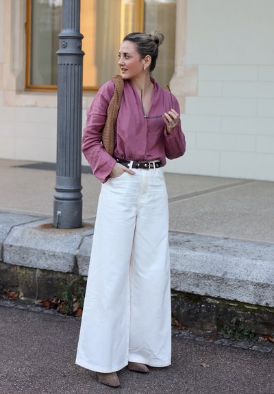 Mujer con una blusa morada y pantalones blancos de pierna ancha, de pie en una acera, sosteniendo unas gafas de sol y un bolso marrón sobre su hombro.