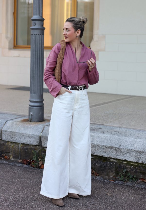 Donna con una blusa viola e pantaloni bianchi a gamba larga, in piedi su un marciapiede, tiene occhiali da sole e una borsa marrone sulla spalla.