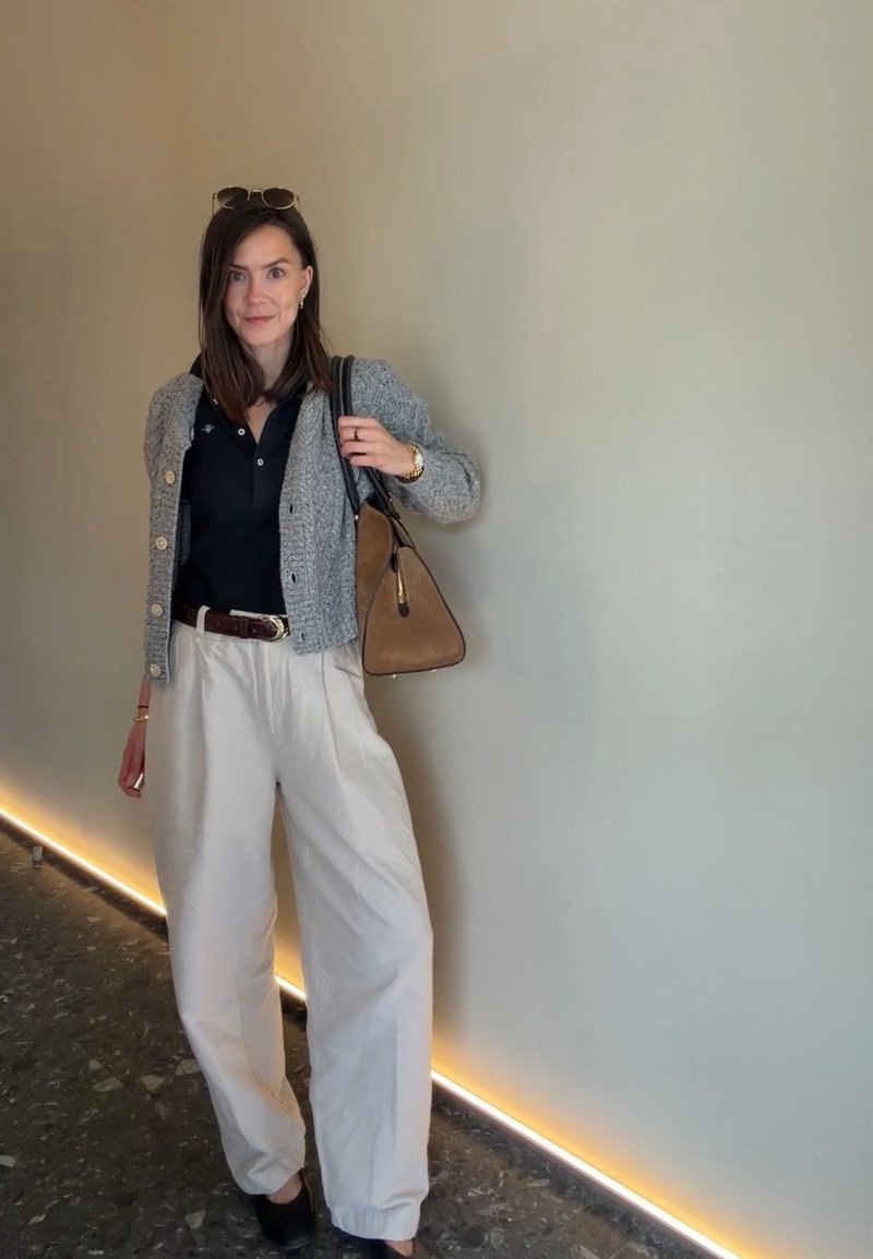 Woman standing indoors wearing a grey cardigan, black shirt, white trousers, black shoes, sunglasses on head, and carrying a brown handbag over one shoulder.