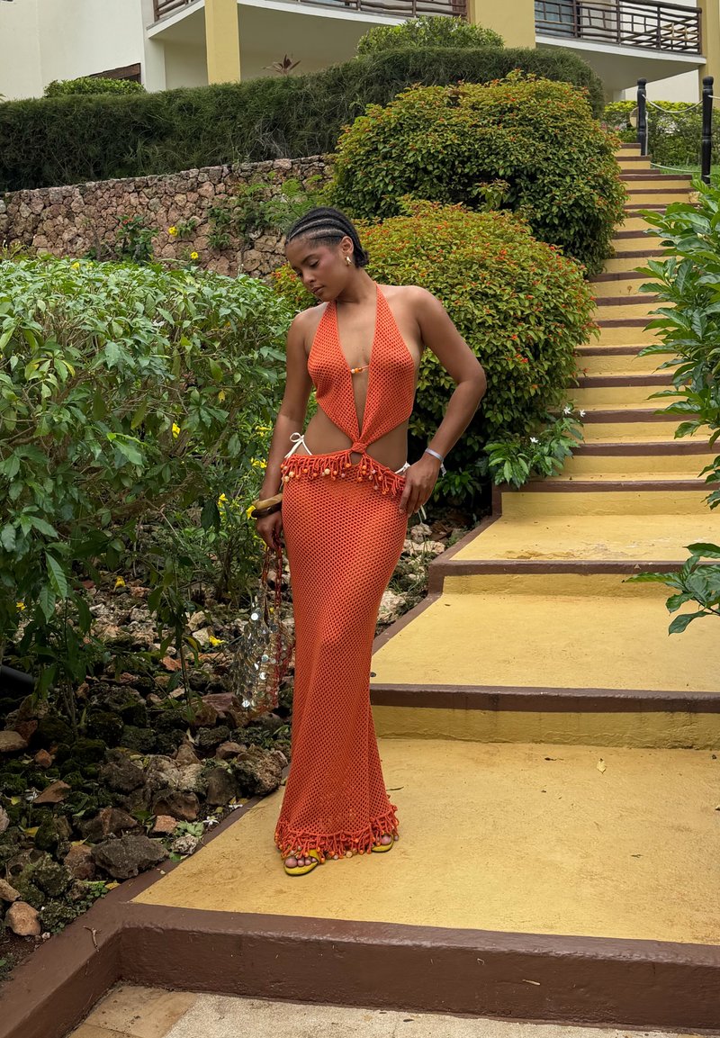 Femme portant une robe à bretelles en crochet orange, debout sur un passage jaune près de plantes vertes et d'escaliers, tenant un sac transparent avec des décorations argentées.