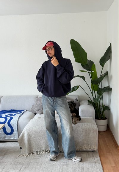 Hombre con sudadera negra con capucha, gorra roja, gafas y vaqueros está en la sala cerca de un sofá con manta gris y una planta grande en maceta.