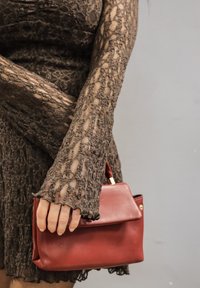 Pruun pitskleit pikkade varrukatega ja lõhenevate mansettidega; hoiab käes väikest, struktureeritud punast nahast käekotti, millel on ülemine käepide ja kuldsed detailid.