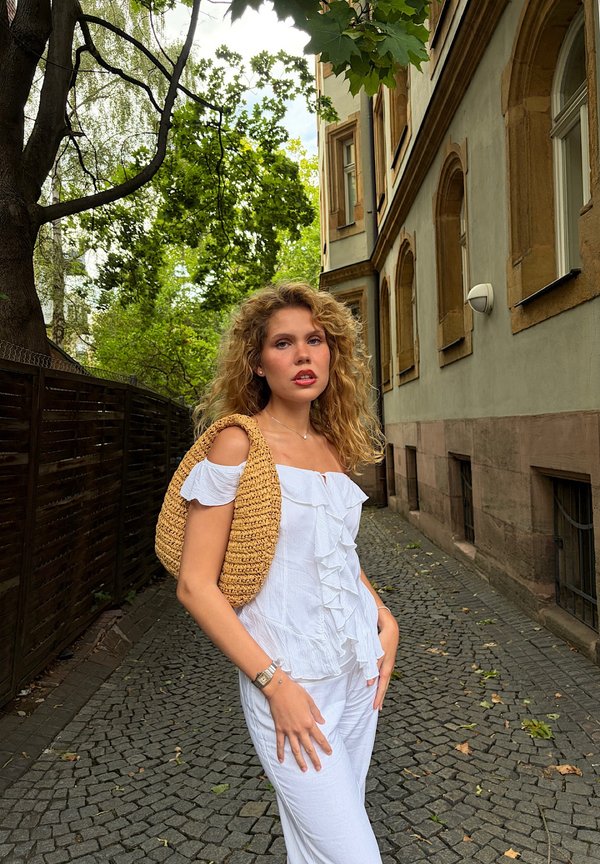 Young woman with curly hair wearing white ruffled top and pants, carrying a woven shoulder bag, standing on a cobblestone street near buildings.