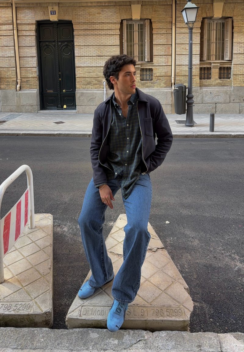 Jeune homme en veste bleue, chemise à carreaux et jean assis sur un bloc de béton triangulaire dans une rue en ville près de l'entrée d'un bâtiment.