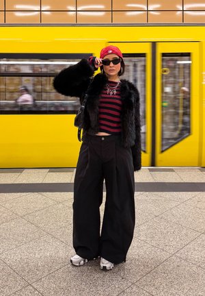 Modelo con abrigo de piel sintética negra, top corto a rayas rojas, pantalones anchos negros y zapatillas plateadas. Sosteniendo gafas de sol frente a un tren amarillo.