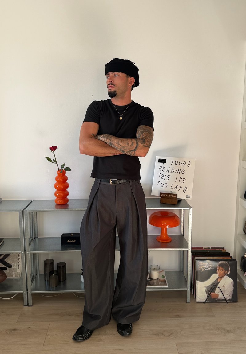 Homme avec un bras tatoué, chemise noire et pantalon gris ample, debout les bras croisés dans une pièce minimaliste avec des étagères portant une rose, une lampe et des disques vinyles.
