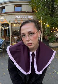 Modell sötét kabátot visel nagy, lila gallérral és rózsaszín szegéllyel, kerek szemüveggel és ezüst fülbevalókkal, kávézóban, kültéri ülőhelyekkel.