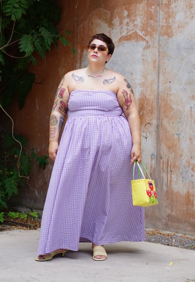 Vestido maxi de cuadros morados con un cuerpo ajustado y una falda fluida, combinado con un pequeño bolso de mimbre amarillo y sandalias deslizantes color tostado.