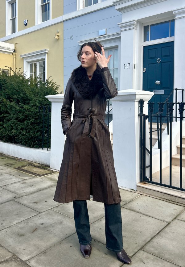 Donna che indossa un lungo cappotto di pelle marrone scuro con colletto di pelliccia, in piedi sul marciapiede davanti a case a schiera dai colori pastello con porta blu.