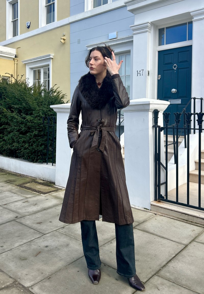 Femme portant un long manteau en cuir marron foncé avec un col en fourrure, debout sur le trottoir devant des maisons de ville aux couleurs pastel avec une porte bleue.