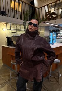 Ruskea nahkatakki, jossa on korkea kaulus, pehmeä koostumus, epäsymmetrinen muotoilu ja kerroksellinen helma. Päällä mustat aurinkolasit. Taustalla baarimiljöö.