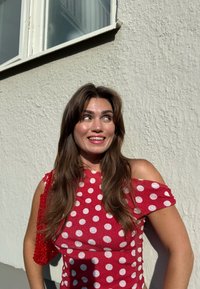 Jovem mulher com um vestido vermelho e branco de bolinhas, sorrindo e olhando para cima, de pé contra uma parede branca texturizada com uma janela acima.