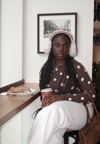 Hnědý top s dlouhým rukávem s puntíky, bílé široké kalhoty, bílé koženkové náušníky. Drží šálek kávy vedle pečiva na dřevěném stole.