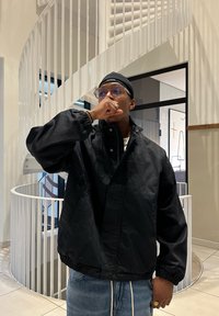 Mann i svart jakke, briller og hodeplagg står innendørs foran en hvit spiraltrapp, og dekker munnen med hånden.